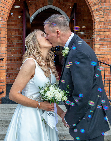 a photo of a bride and groom kissing in front of a brick church with bubbles flying around them. Photo taken by Rossi photo company in Orange Beach, Alabama. Professional photographer in Orange Beach Alabama.